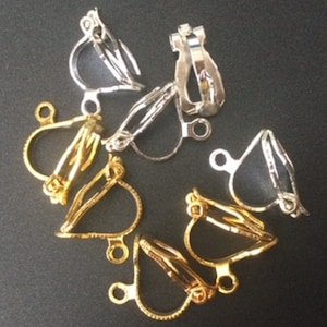 May include: A selection of clip-on earring findings in gold and silver hues. The findings feature a hinged design with a small loop for attaching decorative elements. The metal pieces are arranged on a dark surface.