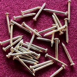 May include: A pile of gold-coloured tacks or upholstery nails on a burgundy surface. The tacks are scattered randomly and some are overlapping.