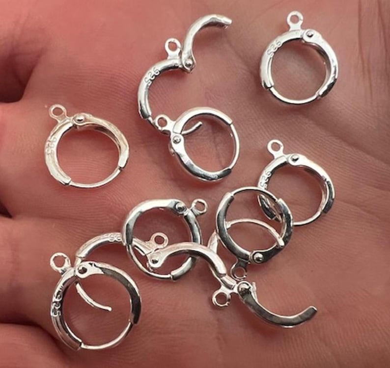 May include: A close-up of a group of silver earring hoops with a clasp closure. The hoops are arranged in a random pattern on a hand.