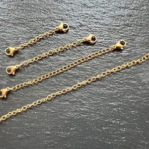 May include: Five gold-toned extender chains with lobster claw clasps. The chains are arranged in a criss-cross pattern on a dark grey surface.