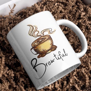 May include: A white ceramic mug featuring a graphic illustration of a steaming cup of coffee in shades of brown and yellow. Below the illustration, the word "Brew-tiful" is printed in a black cursive script font on the side of the mug.