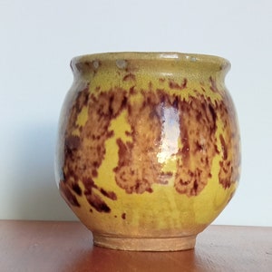 May include: A small, round, yellow ceramic pot with brown and yellow glaze. The pot has a crackled glaze and a slightly uneven surface.
