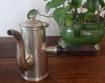 Op de afbeelding: Een zilveren koffiekan met een houten handvat. De kan heeft een rond lichaam en een tuit die naar boven buigt. Het handvat is aan de zijkant van de kan bevestigd en is gemaakt van donker hout.