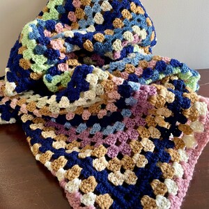 K&ouml;nnte beinhalten: Eine geh&auml;kelte Decke mit einem Granny-Square-Muster in Blaut&ouml;nen, Gr&uuml;n, Rosa, Braun und Wei&szlig;.