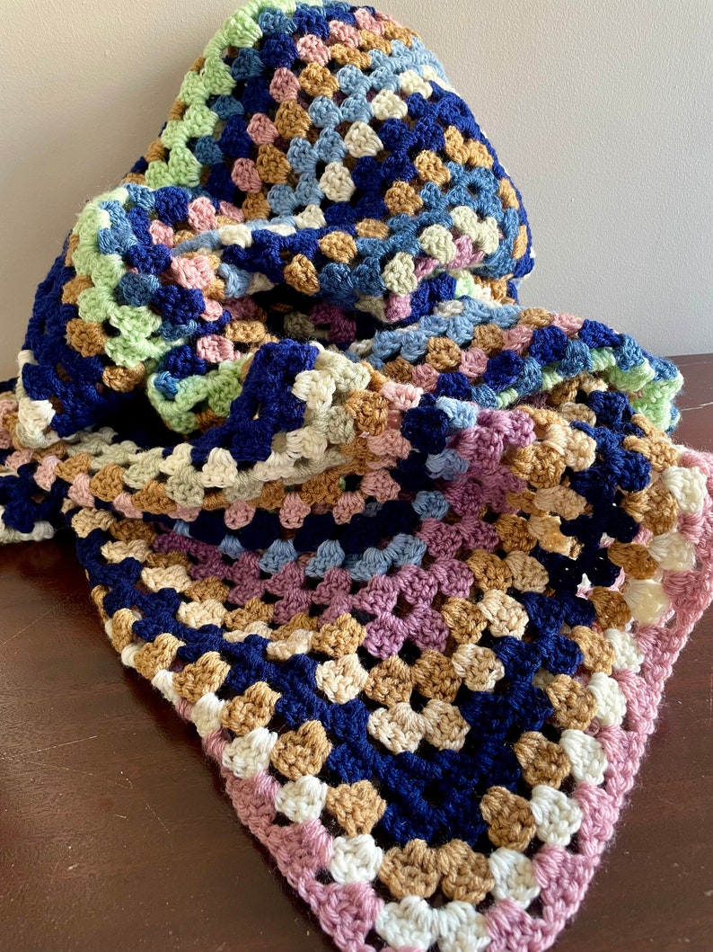 K&ouml;nnte beinhalten: Eine geh&auml;kelte Decke mit einem Granny-Square-Muster in Blaut&ouml;nen, Gr&uuml;n, Rosa, Braun und Wei&szlig;.