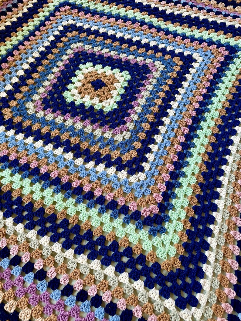 K&ouml;nnte beinhalten: Eine geh&auml;kelte Decke mit einem quadratischen Muster in Blaut&ouml;nen, Gr&uuml;n, Braun, Wei&szlig; und Rosa.