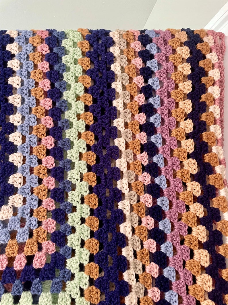 K&ouml;nnte beinhalten: Eine mehrfarbige geh&auml;kelte Decke mit einem diagonalen Muster in Blaut&ouml;nen, Gr&uuml;n, Rosa, Braun und Beige.
