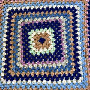K&ouml;nnte beinhalten: Eine quadratische H&auml;keldecke mit einem mehrfarbigen Granny-Square-Muster in Blaut&ouml;nen, Gr&uuml;n, Braun und Rosa.