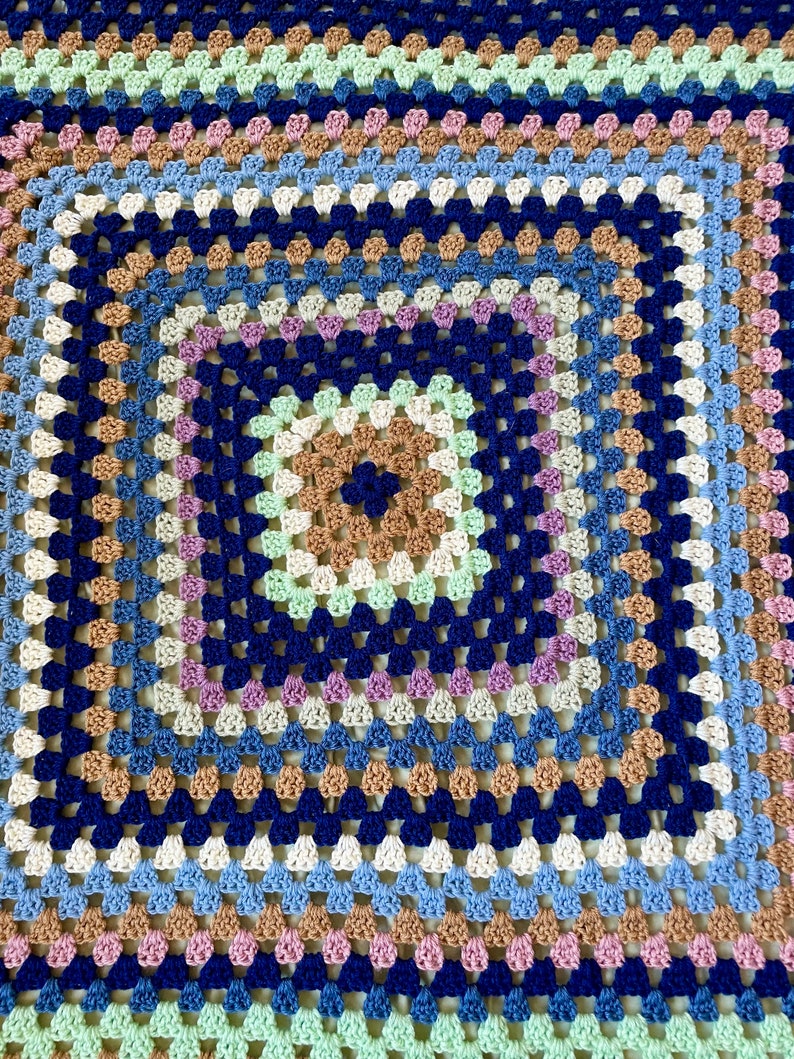 K&ouml;nnte beinhalten: Eine quadratische H&auml;keldecke mit einem mehrfarbigen Granny-Square-Muster in Blaut&ouml;nen, Gr&uuml;n, Braun und Rosa.