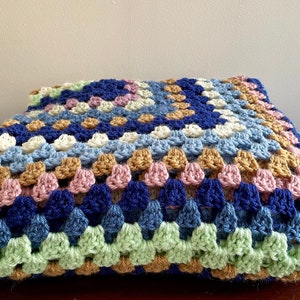 K&ouml;nnte beinhalten: Eine geh&auml;kelte Wolldecke mit einem Granny-Square-Muster in Blaut&ouml;nen, Gr&uuml;n, Braun, Rosa und Wei&szlig;.