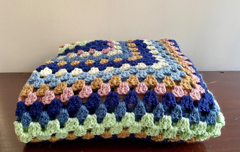 K&ouml;nnte beinhalten: Eine geh&auml;kelte Wolldecke mit einem Granny-Square-Muster in Blaut&ouml;nen, Gr&uuml;n, Braun, Rosa und Wei&szlig;.