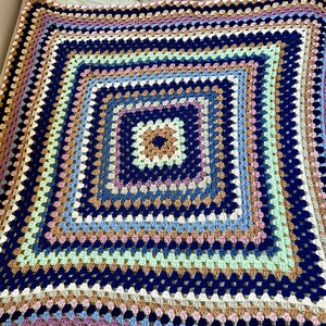 K&ouml;nnte beinhalten: Eine gro&szlig;e geh&auml;kelte Decke mit einem quadratischen Muster in Blaut&ouml;nen, Braun, Beige, Gr&uuml;n und Lila. Die Decke ist mit einem Granny-Square-Stich gearbeitet und hat eine strukturierte, handgefertigte Optik.