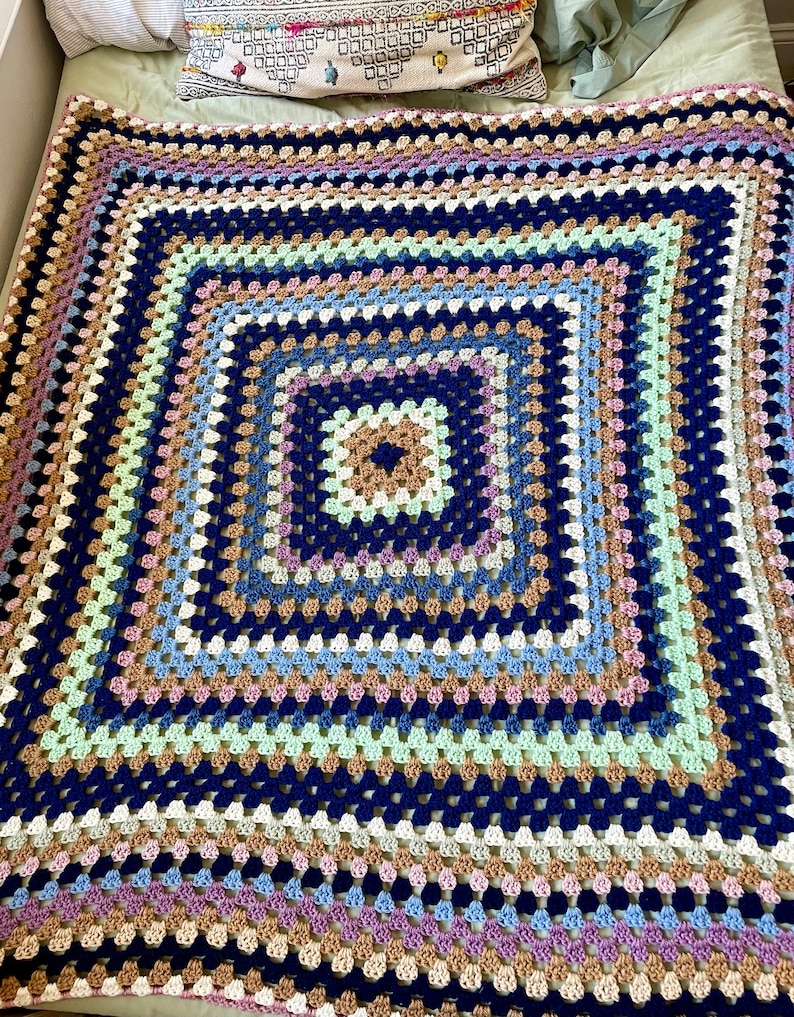 K&ouml;nnte beinhalten: Eine gro&szlig;e geh&auml;kelte Decke mit einem quadratischen Muster in Blaut&ouml;nen, Braun, Beige, Gr&uuml;n und Lila. Die Decke ist mit einem Granny-Square-Stich gearbeitet und hat eine strukturierte, handgefertigte Optik.