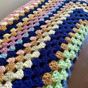K&ouml;nnte beinhalten: Eine geh&auml;kelte Decke mit einem Granny-Square-Muster in den Farben Rosa, Blau, Gr&uuml;n, Beige und Dunkelblau.
