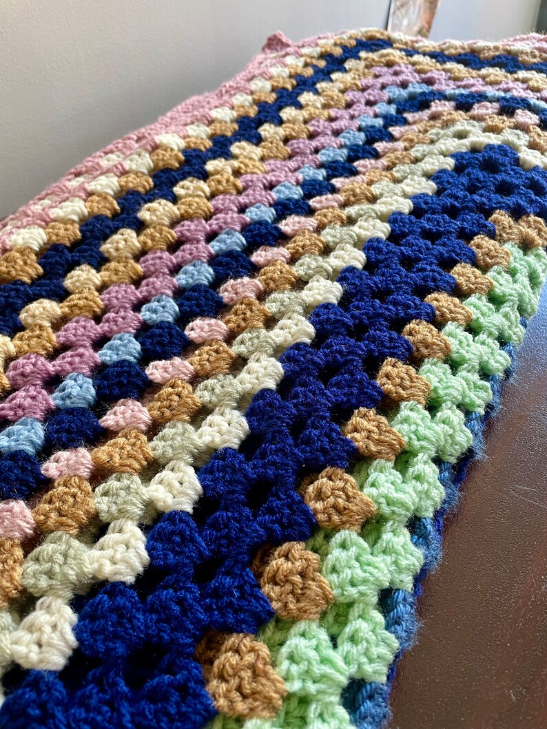 K&ouml;nnte beinhalten: Eine geh&auml;kelte Decke mit einem Granny-Square-Muster in den Farben Rosa, Blau, Gr&uuml;n, Beige und Dunkelblau.