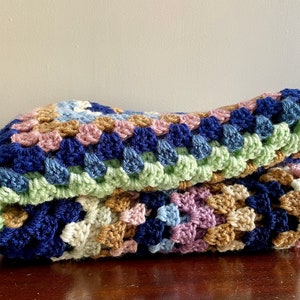 K&ouml;nnte beinhalten: Eine geh&auml;kelte Decke mit einem Granny-Square-Muster in Blaut&ouml;nen, Gr&uuml;n, Rosa und Braun.