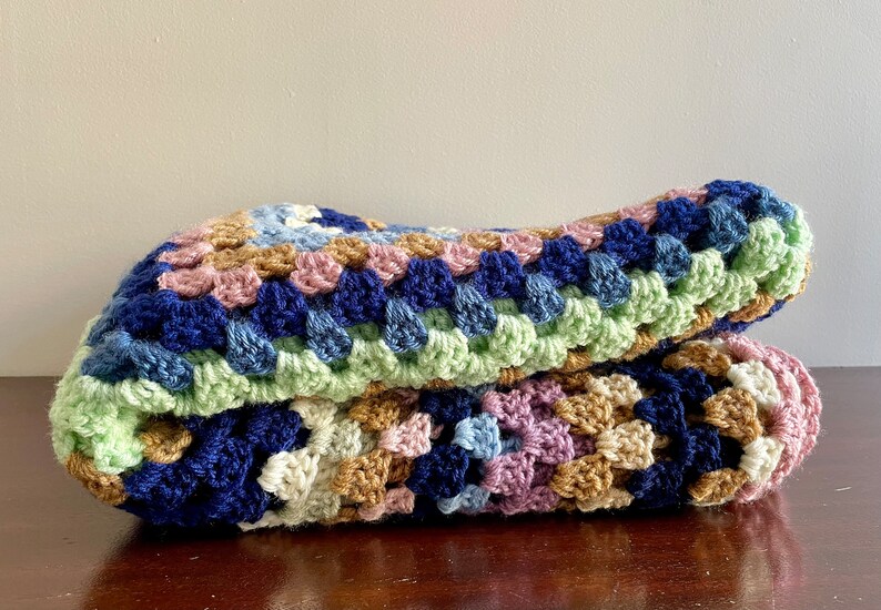K&ouml;nnte beinhalten: Eine geh&auml;kelte Decke mit einem Granny-Square-Muster in Blaut&ouml;nen, Gr&uuml;n, Rosa und Braun.