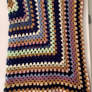 K&ouml;nnte beinhalten: Eine mehrfarbige geh&auml;kelte Decke mit einem Granny-Square-Muster. Die Decke ist aus verschiedenen Farben gefertigt, darunter Rosa, Blau, Gr&uuml;n, Braun und Wei&szlig;.