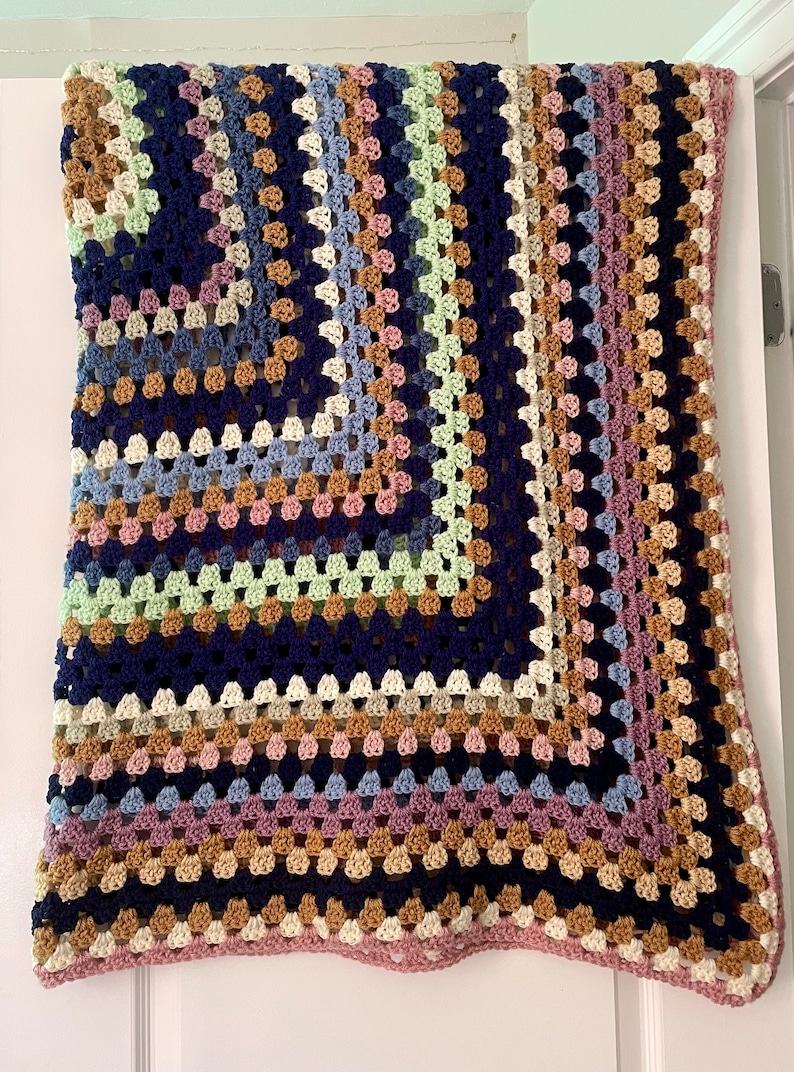 K&ouml;nnte beinhalten: Eine mehrfarbige geh&auml;kelte Decke mit einem Granny-Square-Muster. Die Decke ist aus verschiedenen Farben gefertigt, darunter Rosa, Blau, Gr&uuml;n, Braun und Wei&szlig;.