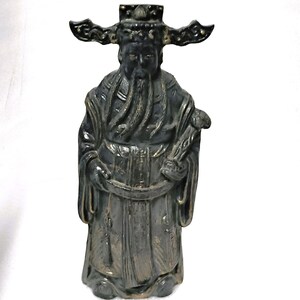 May include: A dark gray statue of a robed figure with a long beard and a hat with wing-like accents. The figure is holding a scroll and has intricate details in the clothing and facial features. The statue is on a white background.