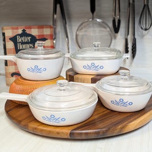 May include: A set of four white ceramic cookware pieces with blue floral patterns and clear glass lids. The collection includes two saucepans with handles and two rectangular or round dishes, all featuring matching blue botanical designs.