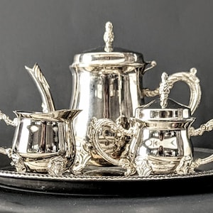 May include: A silver tea set with ornate handles and detailing, including a teapot, creamer, sugar bowl, and spoon, presented on a round tray. The set has a reflective surface, and the background is dark.