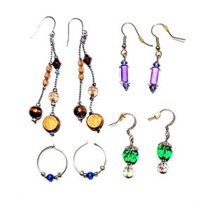 May include: A collection of handmade earrings in various styles. The earrings feature a variety of beads in brown, gold, purple, green, and blue. Some earrings have a dangling design, while others are hoops.