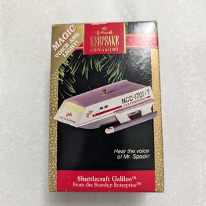 May include: A Hallmark Keepsake Ornament box featuring the Star Trek Shuttlecraft Galileo. The box is gold, black, and red with text reading Magic Voice and Light, NCC-1701/7, U.S.S. Enterprise, Hear the voice of Mr. Spock, and Shuttlecraft Galileo.