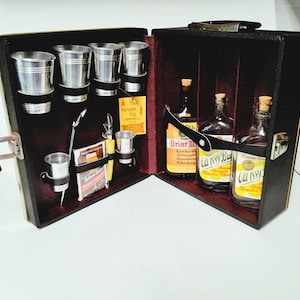 May include: A black portable bar case with a dark red interior, containing three glass whiskey bottles with cork stoppers, five silver-toned metal cups, a metal stirring spoon, a bottle opener, and a yellow tag labeled The Portable Pub.