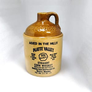 May include: A white ceramic mug with a black handle and the text "But First, Coffee" in a bold, black font. The mug is set against a blurred background, suggesting a cozy setting.