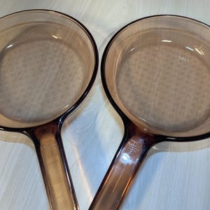 May include: Two matching round skillets made of translucent amber-colored glass. Each skillet features a long, integrated handle with the embossed text Vision Corning USA visible on the neck. The glass has a smooth, polished finish throughout.