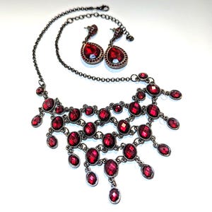 May include: A dark metal necklace and matching earrings set. The necklace features multiple tiers of large, oval, and round red gemstones. The earrings have a teardrop shape with a large red gemstone. The jewelry set is on a white background.