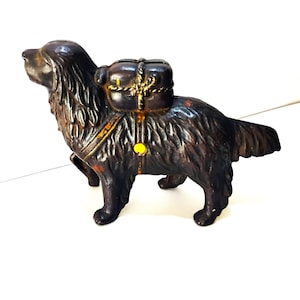 May include: A cast iron dog figurine in a standing pose, with a pack on its back. The dog is dark brown with a textured coat, and the pack is secured with gold-colored rope. A yellow accent is visible on the side.