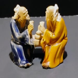 May include: A ceramic figurine depicting two elderly figures seated across from each other. One figure wears a dark blue robe, and the other wears a mustard yellow robe. Both figures have white hair and beards, and they hold small cups near a central vessel.