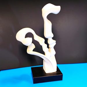 May include: A white abstract sculpture with curved, flowing forms. The sculpture is mounted on a black rectangular base. The background is a gradient of black and blue.