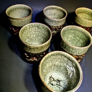 May include: Six ceramic tea cups with a crackle glaze in shades of green and brown. The cups have a dark brown base with a decorative pattern. One cup features a black horse illustration inside.