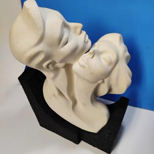 May include: A cream-colored sculpture depicting two faces in profile, leaning towards each other. The sculpture is mounted on a black, geometric base. The faces have closed eyes and smooth features, suggesting a theme of intimacy or connection.