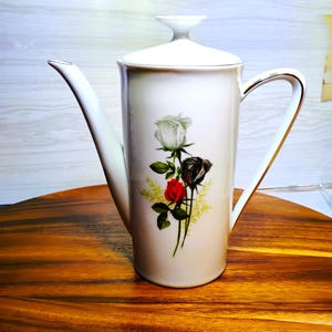 May include: White ceramic coffee pot with a lid, a long spout, and a gold handle. The pot is decorated with a floral design featuring a white, black, and red rose bouquet. The pot is sitting on a wooden surface.
