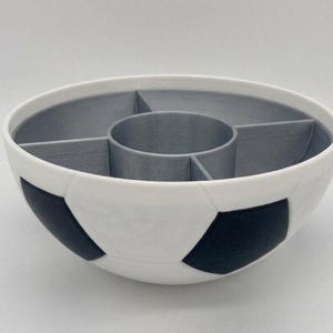 May include: A round bowl designed with a soccer ball pattern featuring white and black exterior panels. The interior is divided into four outer sections and one central circular compartment, all finished in a solid matte grey color.
