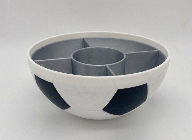 May include: A round bowl designed with a soccer ball pattern featuring white and black exterior panels. The interior is divided into four outer sections and one central circular compartment, all finished in a solid matte grey color.