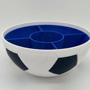 May include: A white bowl with a black soccer ball pattern on the exterior. The interior is blue and features a central circular compartment surrounded by four separate wedge-shaped sections, creating a divided container for organizing small items.