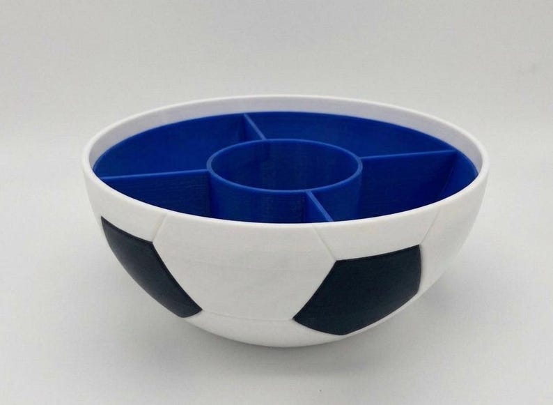 May include: A white bowl with a black soccer ball pattern on the exterior. The interior is blue and features a central circular compartment surrounded by four separate wedge-shaped sections, creating a divided container for organizing small items.