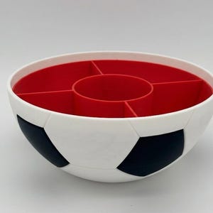 May include: A round, white bowl with a black pentagonal soccer ball pattern on the exterior. The interior is bright red and features a central circular compartment surrounded by four additional wedge-shaped sections for organized storage.