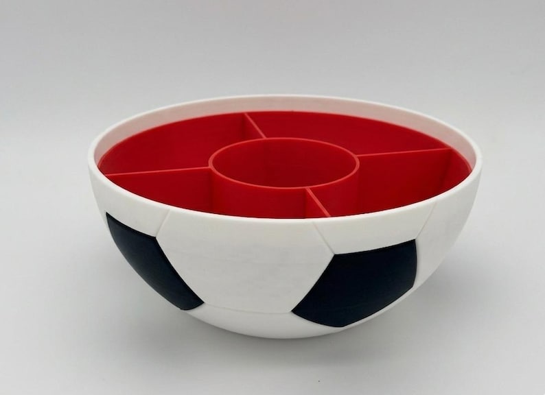 May include: A round, white bowl with a black pentagonal soccer ball pattern on the exterior. The interior is bright red and features a central circular compartment surrounded by four additional wedge-shaped sections for organized storage.