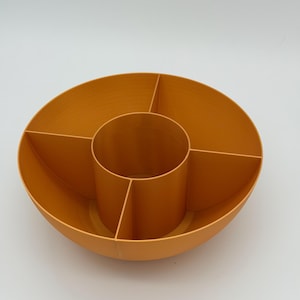 May include: A circular orange storage organizer featuring a central cylindrical compartment surrounded by four distinct wedge-shaped sections. The container has smooth, solid walls and an open top design, providing multiple divided spaces for sorting items.