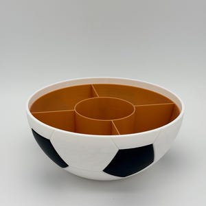 May include: A round, bowl-shaped organizer featuring a white and black exterior patterned like a soccer ball. The interior is a solid tan color and is divided into five distinct sections, including a central circular compartment and four surrounding ones.