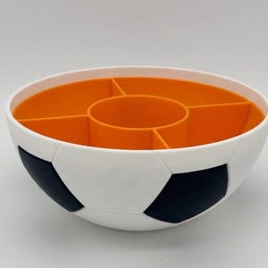 May include: A round bowl designed with a white and black soccer ball pattern on the exterior. The interior is bright orange and features a central circular compartment surrounded by four additional wedge-shaped sections for organized storage.