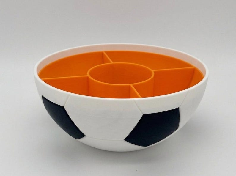 May include: A round bowl designed with a white and black soccer ball pattern on the exterior. The interior is bright orange and features a central circular compartment surrounded by four additional wedge-shaped sections for organized storage.