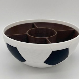 May include: A round serving bowl designed with a white and black soccer ball pattern on the exterior. The interior is dark brown and features a central circular compartment surrounded by four separate wedge-shaped sections for food organization.