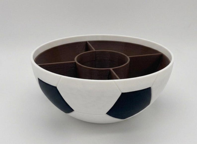 May include: A round serving bowl designed with a white and black soccer ball pattern on the exterior. The interior is dark brown and features a central circular compartment surrounded by four separate wedge-shaped sections for food organization.
